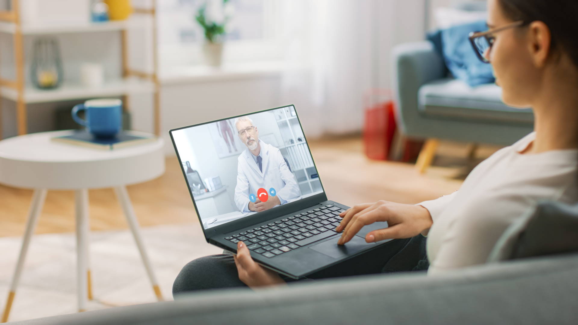 A woman sitting on a couch in a room talks with a doctor providing telemedicine services over a video call.